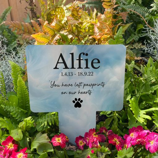 Clouds in heaven background with silhouette of paw print and the text you have left paw prints on our hearts dog memorial stake inserted in flower bed surrounded by colourful flowers in the garden