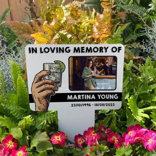 White background with hand raising a glass of gin memorial stake inserted in flower bed surrounded by colourful flowers in the garden