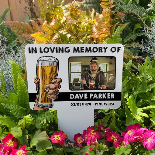 White background with hand raising pint of lager memorial stake inserted in flower bed surrounded by colourful flowers in the garden