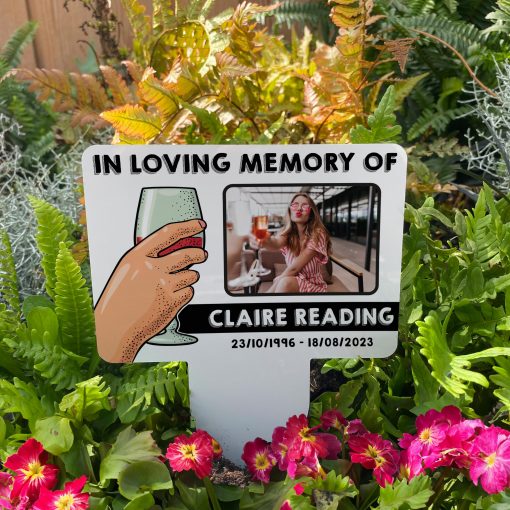 White background with hand raising a glass of red wine memorial stake inserted in flower bed surrounded by colourful flowers in the garden