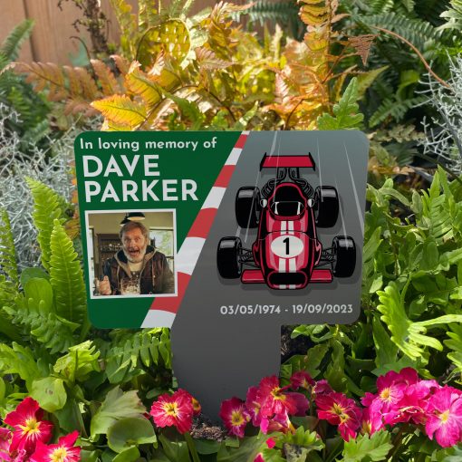 Red retro racing car memorial stake inserted in flower bed surrounded by colourful flowers in the garden