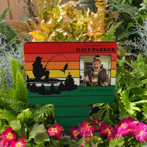 A striped gradient background with a silhouette of a man fishing memorial stake inserted in flower bed surrounded by colourful flowers in the garden