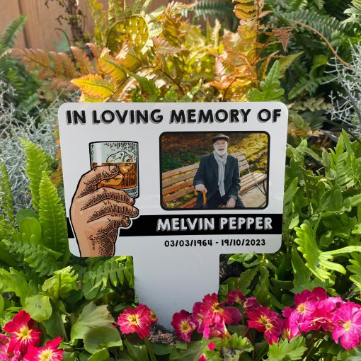 White background with hand raising a glass of whiskey memorial stake inserted in flower bed surrounded by colourful flowers in the garden